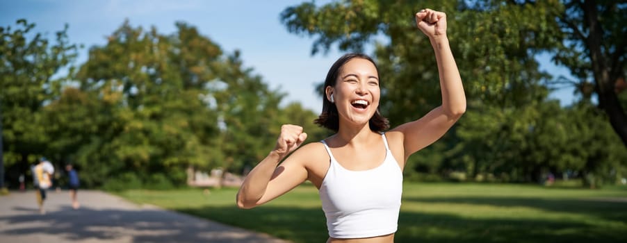 Happy fitness girl achieve goal, finish marathon, running with hands up, celebrating victory while jogging, triumphing in park.