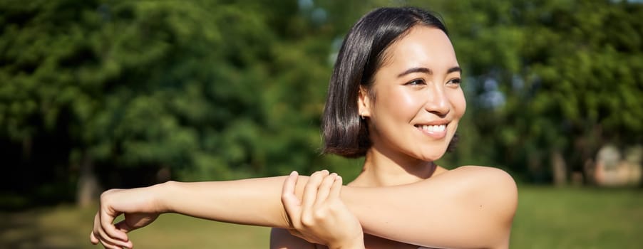 Portrait of young fitness woman stretching her arms, warm-up before training session, sport event in park, jogging and excercising.