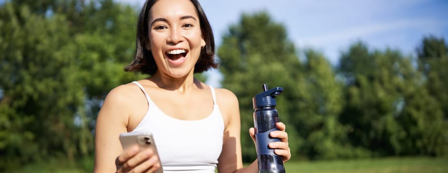 Excited young asian woman workout and drinks water, follows sports app on smartphone, looks amazed and happy, does excercises in park.