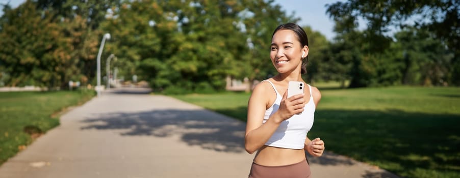 Happy smiling asian woman jogging in park. Healthy young female runner doing workout outdoors, running on streets.