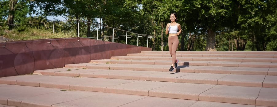 Portrait of young sportswoman running on street. Fitness girl jogging in park, workout outdoors, listening music in wireless earphones.