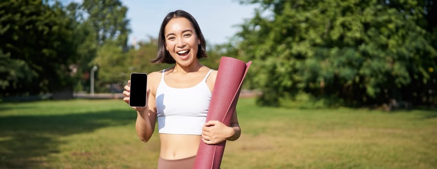 Smiling asian fitness girl with rubber yoga mat, shows her smartphone screen, recommends workout application, stands on lawn in park.