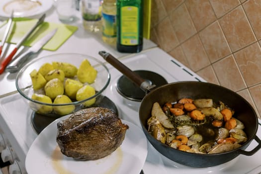 Roast beef lies on a plate next to a pan with vegetables and a bowl of potatoes. High quality photo