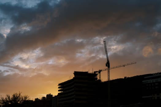 Silhouettes of tower cranes at the construction site of a multi-storey building against the background of an orange sunset sky. High quality photo