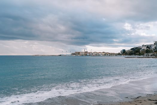 Sea waves roll on the sandy beach against the backdrop of a stormy sky and high-rise buildings on the shore. High quality photo