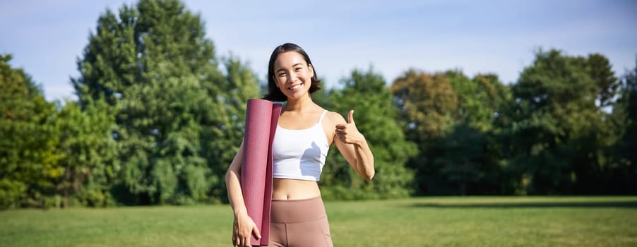 Beautiful and fit fitness girl in leggings, shows thumb up in park, stands with yoga rubber mat for workout and sport training.