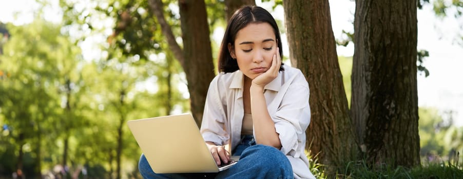 Portrait of asian woman sitting on grass near tree, using laptop, working, doing homework remotely on sunny summer day.