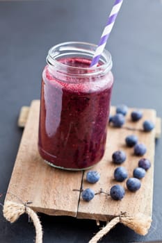 Closeup of blueberry smoothie in glass with striped straw on wooden serving tray with blueberries