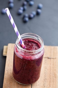 Closeup of blueberry smoothie in glass with striped straw on wooden serving tray with blueberries in background