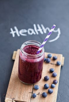 Closeup of blueberry smoothie in glass with striped straw on wooden serving tray with word healthy written on dark background