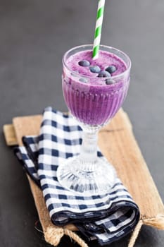 Closeup of blueberry milk smoothie in glass with striped straw on checkered napkin on wooden serving tray