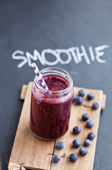 Closeup of blueberry smoothie in glass with striped straw on wooden serving tray with word smoothie written on dark background