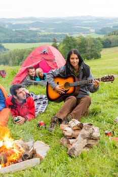 Young woman playing guitar beside camping friends lying in tents