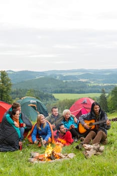 Group of friends sitting beside tents, campfire girl playing guitar