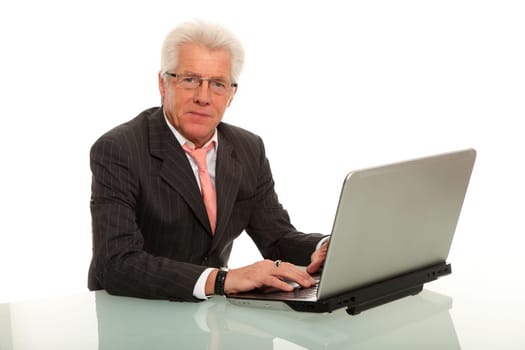 An old man sits at a laptop and looks kindly into the camera.