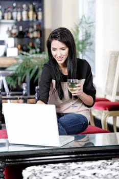 Beautiful young woman chatting with friends on her laptop while enjoying a glass of wine in a bar