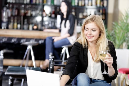 Beautiful young woman chatting with friends on her laptop while enjoying a glass of wine in a bar