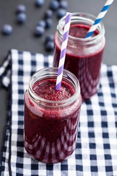 Closeup of two blueberry smoothies in glasses with striped straws resting on checkered napkin with fresh blueberries in background