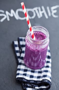 Closeup of blueberry smoothie in glass with striped straw resting on checkered napkin with word smoothie written in chalk on dark background