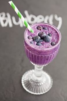 Closeup of blueberry milk smoothie in glass with striped straw and word blueberry written in chalk on dark background