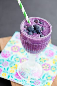 Closeup of blueberry milk smoothie in glass with striped straw on paper serviette on wooden serving tray