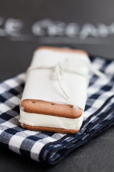 Closeup of one vanilla ice cream biscuit wrapped in white paper with string and bow resting on checkered napkin and word ice cream written in chalk in background