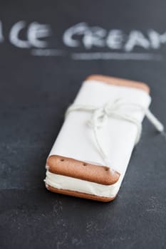 Closeup of one vanilla ice cream biscuit wrapped in white paper with string and bow and word ice cream written in chalk in background
