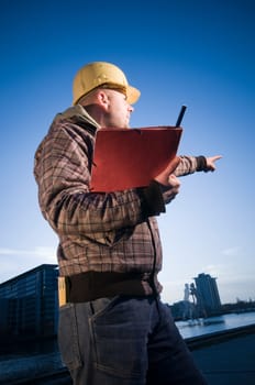 Side three-quarter view of a construction worker with yellow helmet,checkered jacket and red loose-leaf binder with his left arm pointing into the distance and looking in front of evening blue city background