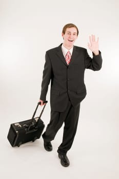 Full-length shot of a young man in a dark suit running towards the camera,pulling a pilot's suitcase behind him in front of a white background,waving at the camera with a friendly smile