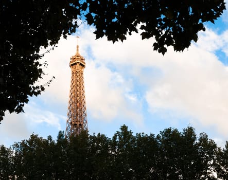 View of top of eiffel tower at sunset. Blue sky with clouds in background. Dark trees in foreground. Paris, France, Europe.