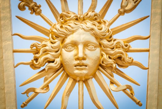 Detail view of golden ornate gate of Chateau de Versailles depicting human face. Blue sky in background. Paris, France, Europe.
