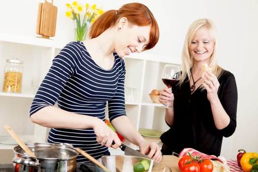 Two attractive women friends preparing a meal in the kitchen together with one chopping vegetables watched by the second who is enjoying a glass of red wine