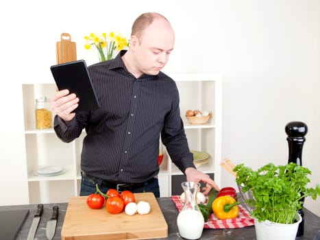 Man cooking in the kitchen carefully checking his fresh ingredients, herb and vegetables while holding a tablet in his other hand