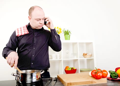 Attractive middle-aged balding man taking a call on his mobile while standing cooking at the stove stirring the contents of a stainless steel saucepan