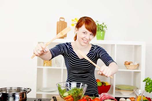 Happy smiling young redhead woman in the kitchen tossing a fresh salad with two wooden salad servers as she prepares a healthy meal