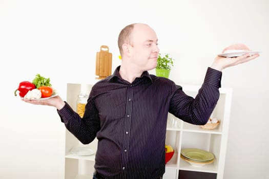 Gleeful smiling man standing in his kitchen with his arms raised with two plates, one with fresh vegetables and one with meat, making a choice on food and diet and giving in to temptation