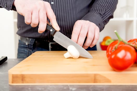 Cropped view of the hands of a man slicing a mushroom and salad ingredients with a large kitchen knife on a wooden chopping board