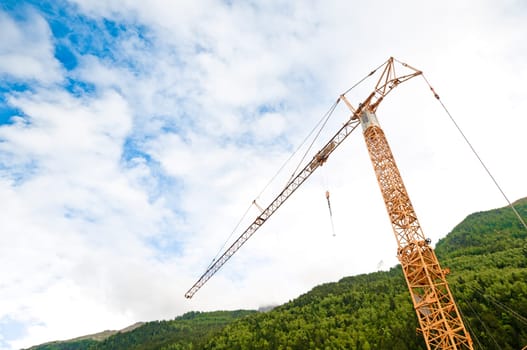 Construction crane on site in Alps mountains. Blue cloudy sky in background and green mountain.
