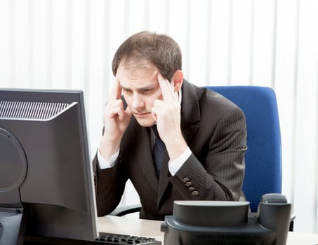 Worried businessman with a headache sitting at his desk rubbing his temples with his fingertips and staring at his desktop computer monitor in despair