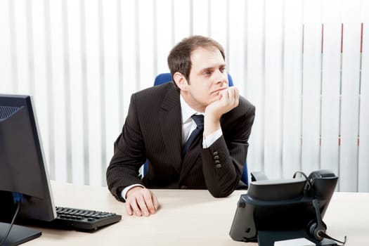 Thoughtful businessman in his office sitting at his desk resting his chin on his hand as he stares into the distance while contemplating difficult decisions