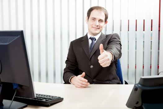 Happy confident businessman sitting at his desk in his office giving a thumbs up gesture of success, approval or to indicate that he has closed a business deal