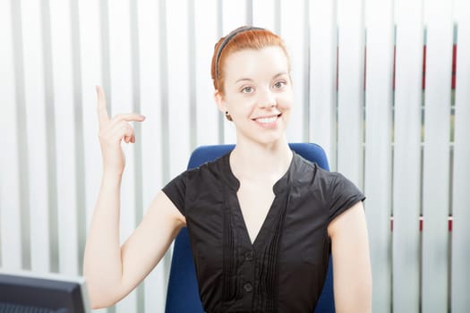 Pretty smart redhead businesswoman with a beautiful smile sitting in her chair at the office pointing above her head
