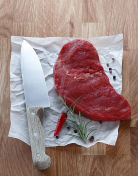 Portion of lean raw steak lying on a crumpled sheet of paper with a chefs knife and fresh rosemary with a red hot chilli pepper in preparation for cooking it