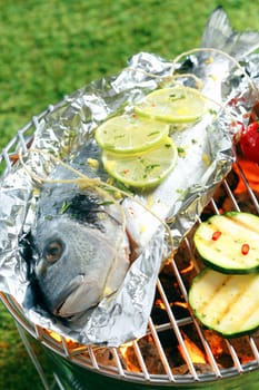 Whole fresh gilthead breem garnished with sliced lemon roasting on aluminium foil over the glowing coals in a barbecue