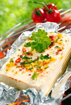 Halloumi cheese topped with fresh herbs and spices grilling in tin foil over the glowing coals of a barbecue outdoors in the sunshine