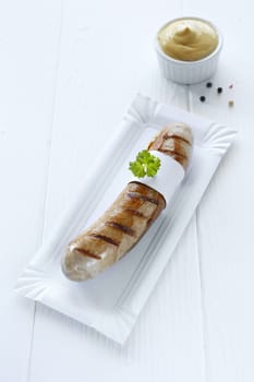 One cooked sausage with a sprig of flat parsley on a white plate with a pot of mustard.