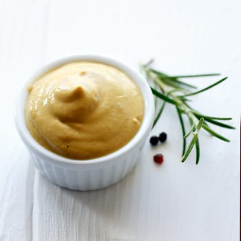 Small ramekin of mustard with a sprig of rosemary against white.