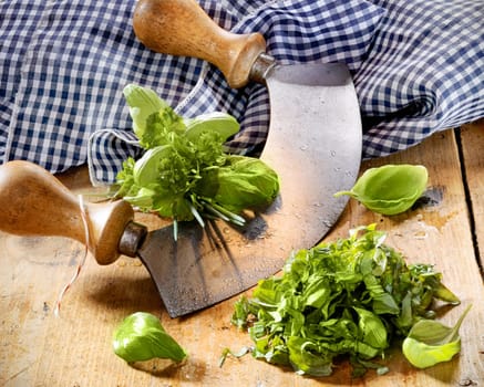 Chopping a selection of mixed herbs in a bunch including basil, parsley and chives using a two handled curved rocker blade on a wooden tabletop