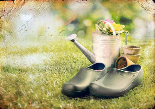 Artistically aged vintage effect image of a pair of green clogs or gardening shoes on the lawn in summer alongside an old metal watering can filled with flowers surrounded by scattered clay flowerpots