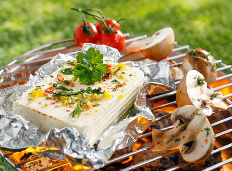 Large portion of seasoned halloumi or feta cheese grilling in tin foil over hot coals in a barbecue outdoors on green grass